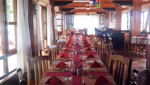 Une longue table à manger, dressée avec des serviettes rouges, des verres et des couverts, entourée de chaises en bois dans une pièce aux poutres apparentes et aux grandes fenêtres.
