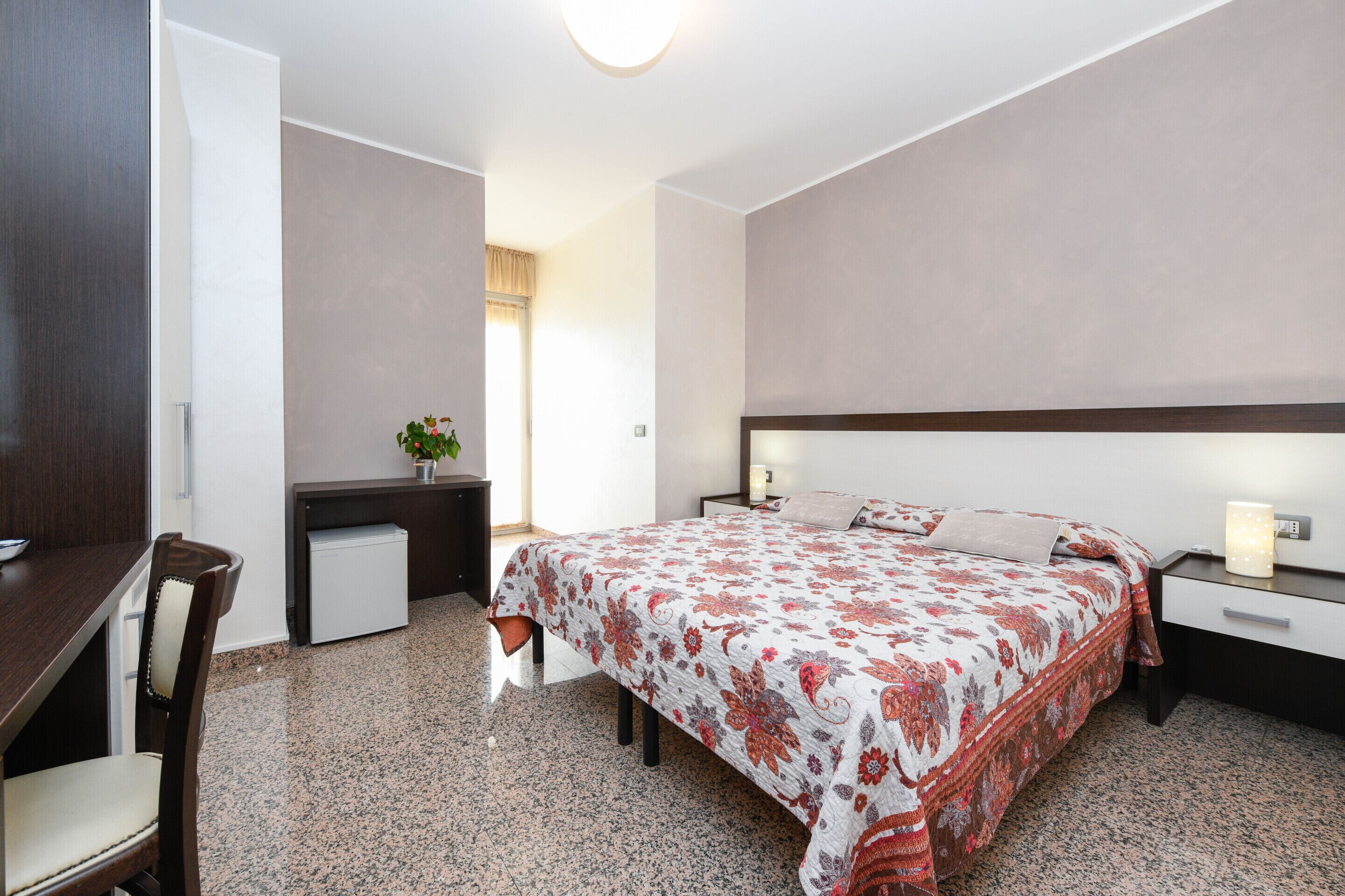 Een slaapkamer met een bed, nachtkastje, bureau, stoel en een kleine koelkast.