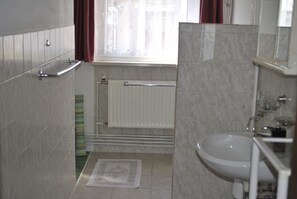 A bathroom with a white sink, a glass shower enclosure, a towel rack, a radiator, and a window with curtains. - Pension Haus Mückenheim (Oberharz am Brocken)