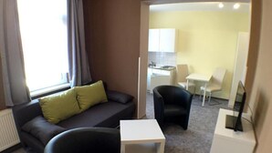 Un espace de vie compact comprenant un canapé, un fauteuil noir, une petite table d’appoint blanche et un téléviseur posé sur un meuble.