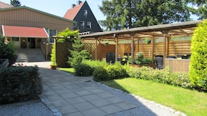 A covered outdoor seating area with tables and chairs, surrounded by a garden with various plants and a gravel path leading to a building entrance. - Pension Haus Mückenheim (Oberharz am Brocken)