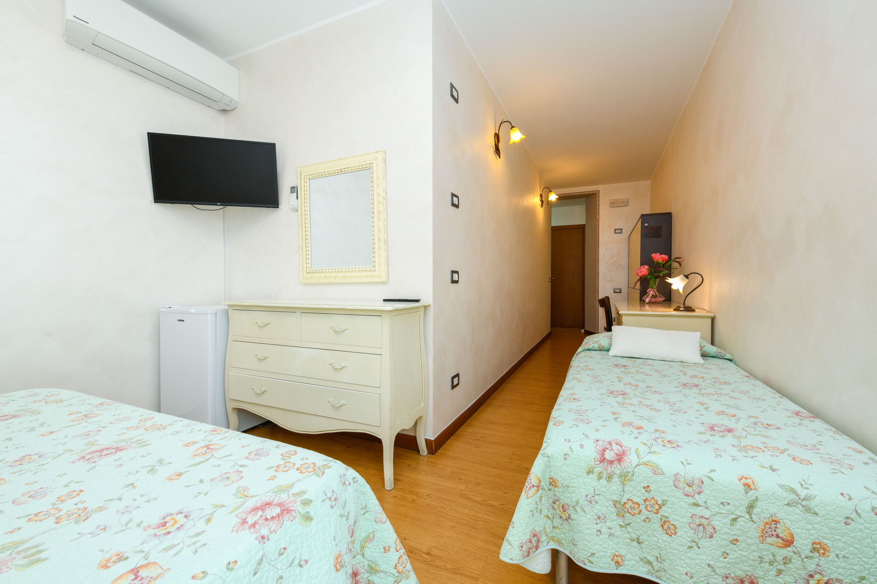 A bedroom with two beds, a TV, a mirror, a dresser, and a small desk with a lamp.