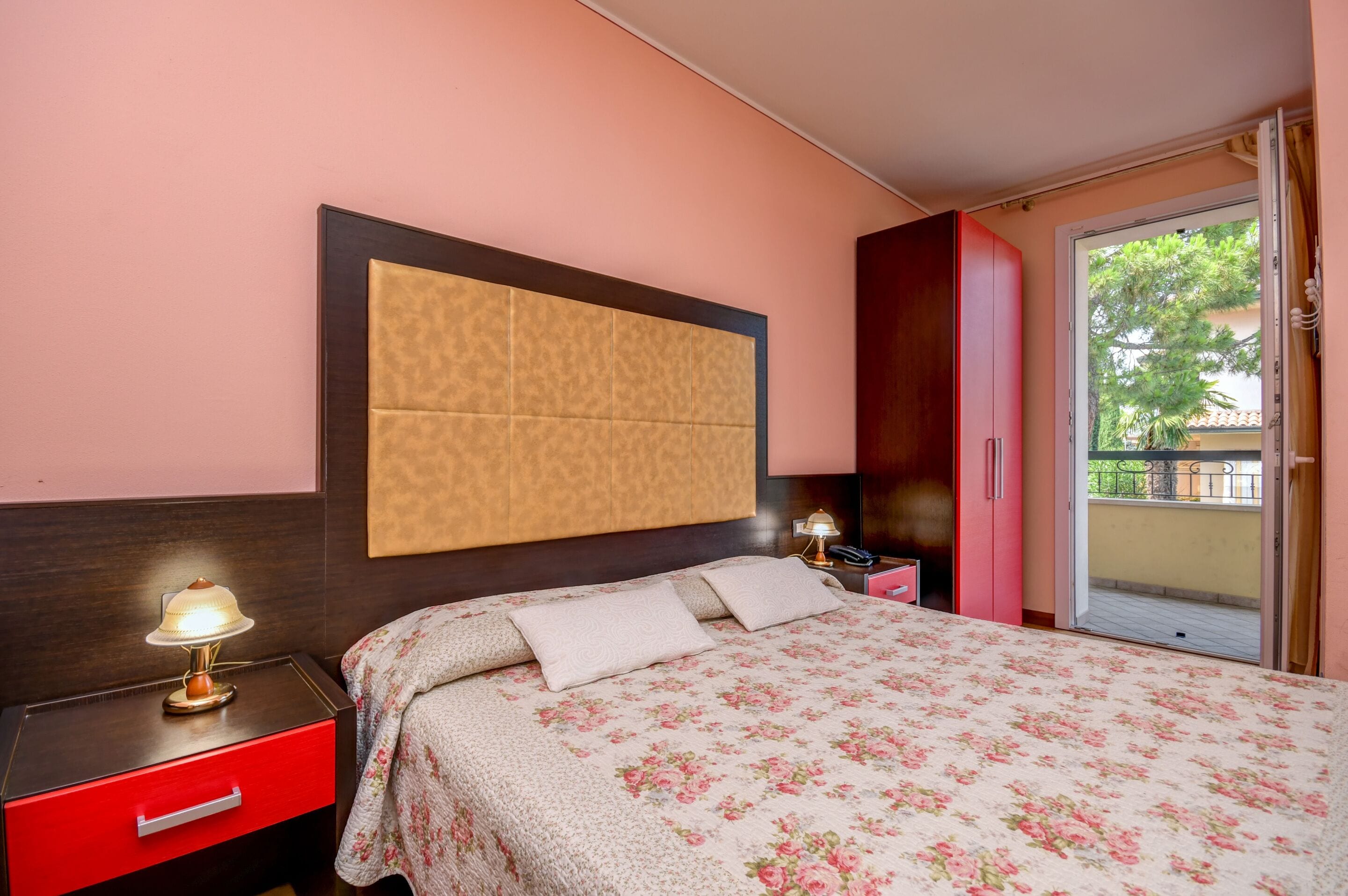 A hotel room with a bed, a nightstand, a red drawer, a lamp, a telephone, a red wardrobe, and a sliding glass door to the outside.