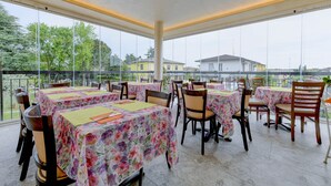 An outdoor dining area with tables and chairs, large windows, and a view of buildings and trees. - B&B America (Sirmione)