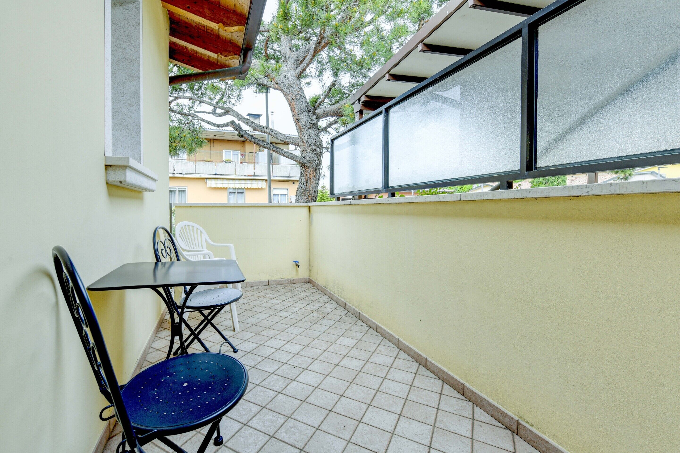 Un pequeño balcón con suelo de baldosas, una mesa, dos sillas y un ventanal grande con vistas a un árbol y edificios.
