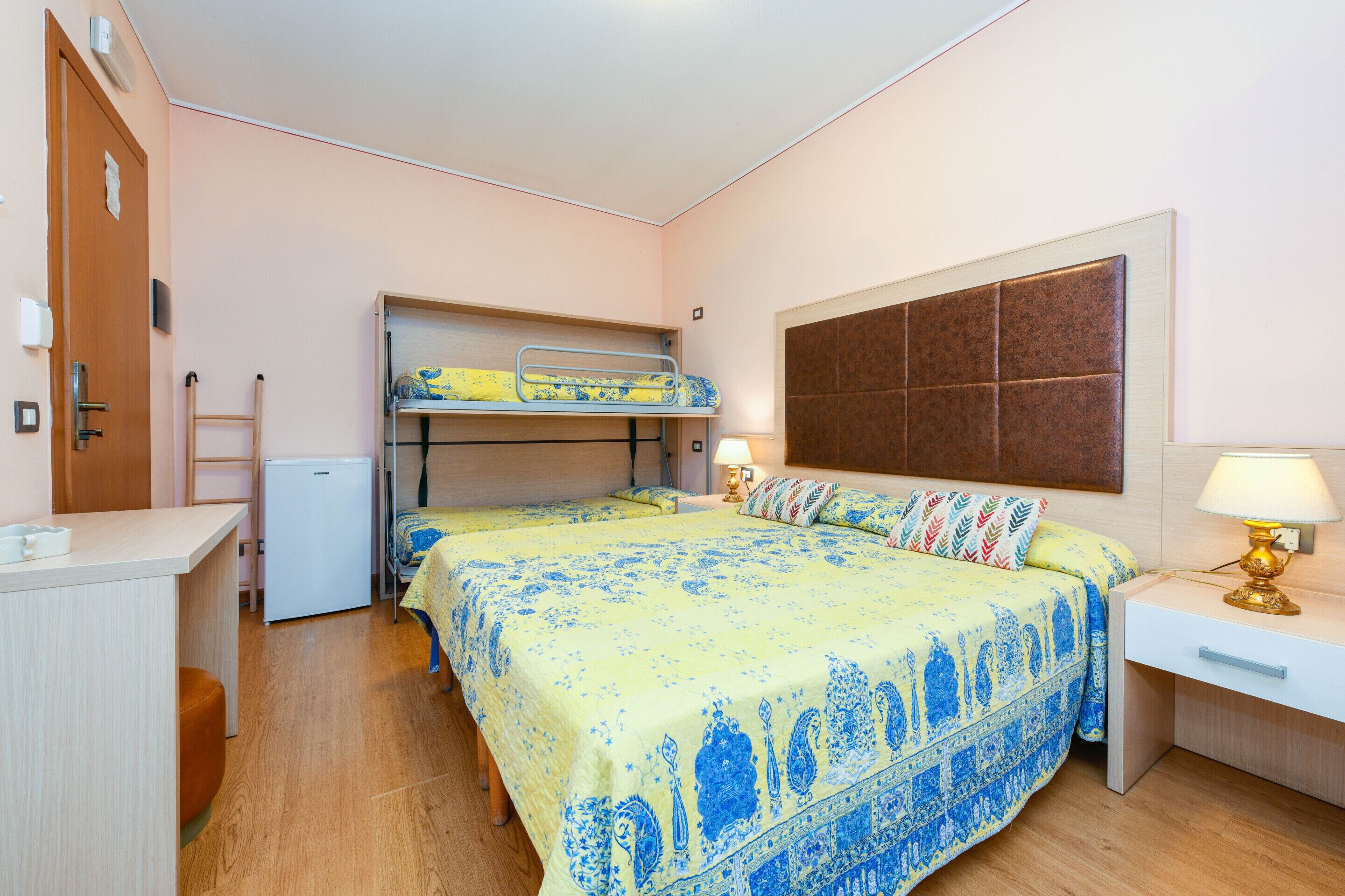 A bedroom with two beds, a desk, a chair, a small fridge, and a bedside lamp.