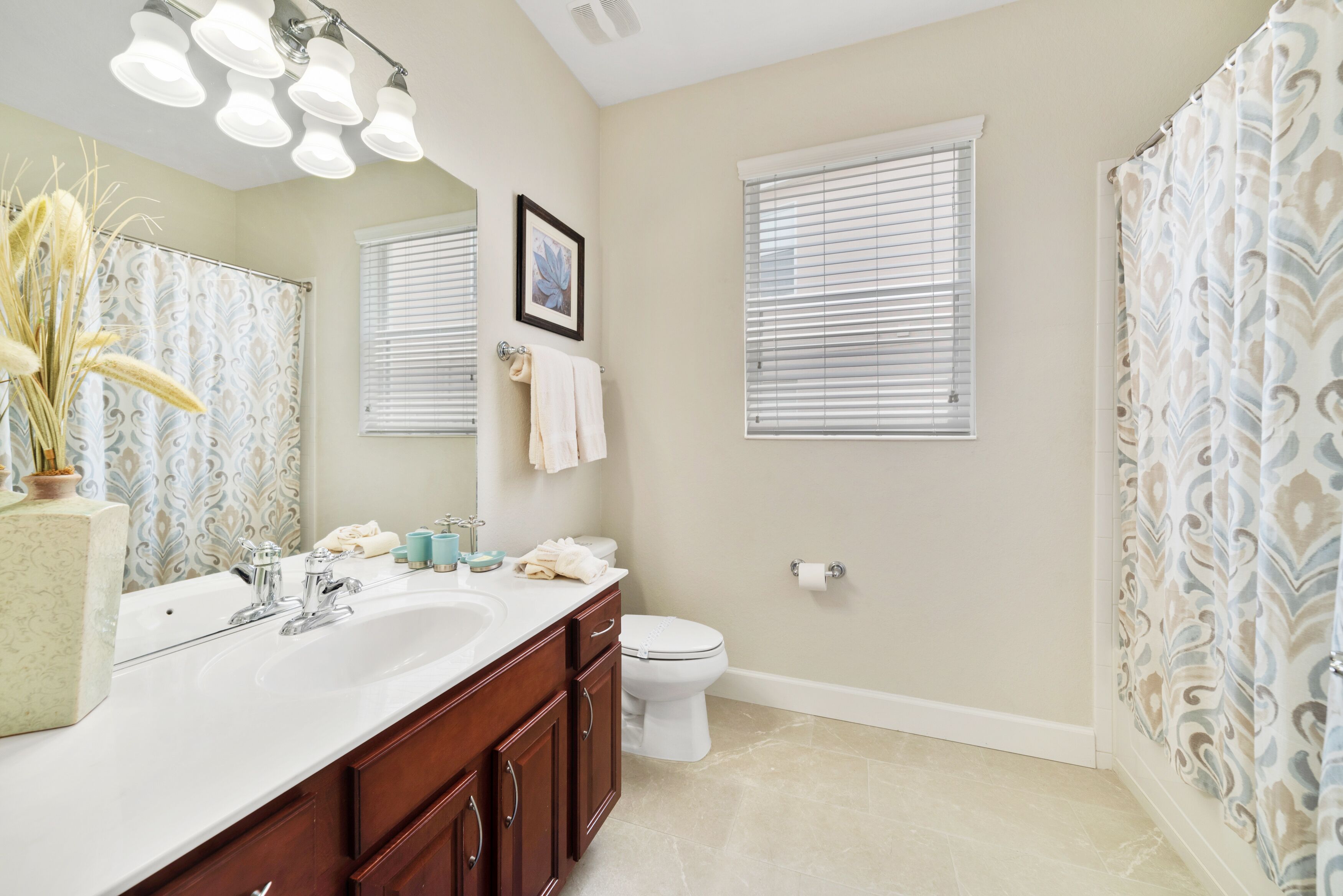 A bathroom with a large sink, vanity, and toilet. There are two windows with blinds, a shower curtain, and a mirror above the sink.