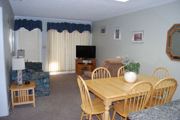 A living room with a television, a sofa, a dining table with chairs, and a potted plant.