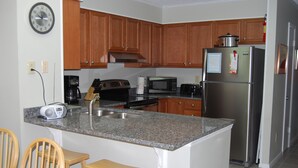 A kitchen with wooden cabinets, a stainless steel refrigerator, a microwave, a coffee maker, and a clock on the wall. - Great View w/o Oceanfront Prices-3BR 2.5BA -  (Myrtle Beach)