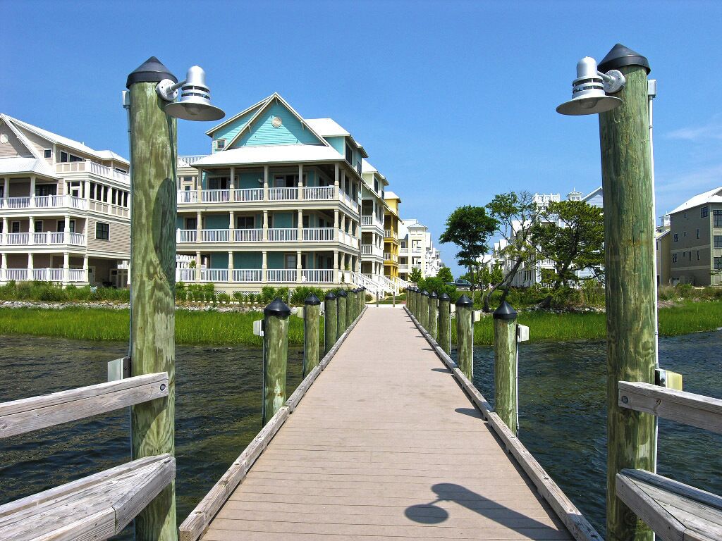 A wooden boardwalk leading to a waterfront property with multiple balconies and a green roof.