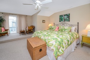 Ein Schlafzimmer mit Bett, Deckenventilator, Fenster mit Vorhängen, Stuhl, Nachttisch mit Lampe und einem Wandbild.