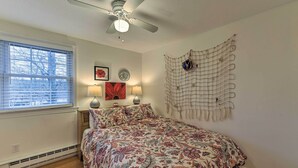Une chambre à coucher comprenant un lit, un ventilateur de plafond, une fenêtre avec des stores et des éléments de décoration murale, notamment un tableau encadré et un élément décoratif suspendu au mur.
