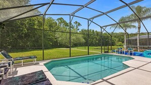 A covered outdoor swimming pool with lounge chairs and a view of a green lawn and trees. - 1061 Lake Berkley 4 Bed with Pool (kissimmee)