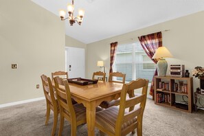 Uma sala de jantar com mesa e cadeiras de madeira, um candelabro, uma estante de livros e uma janela com cortinas.
