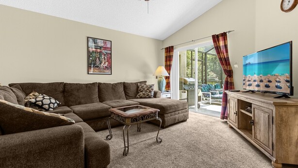 A living room with a sectional sofa, a small table, a TV on a wooden cabinet, and a large window with curtains. - 1061 Lake Berkley 4 Bed with Pool (kissimmee)