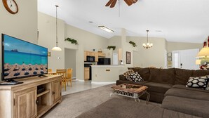 A living room with a large flat-screen TV, a brown sofa, a coffee table, and a dining area with wooden chairs. - 1061 Lake Berkley 4 Bed with Pool (kissimmee)