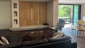 A modern living room with a wooden accent wall, a glass coffee table, a leather sofa, and a balcony with outdoor furniture.