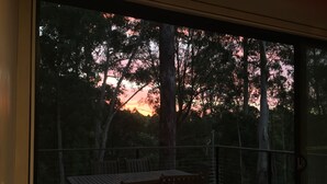 A balcony with a wooden table and chairs, overlooking a forested area with trees and a sunset.