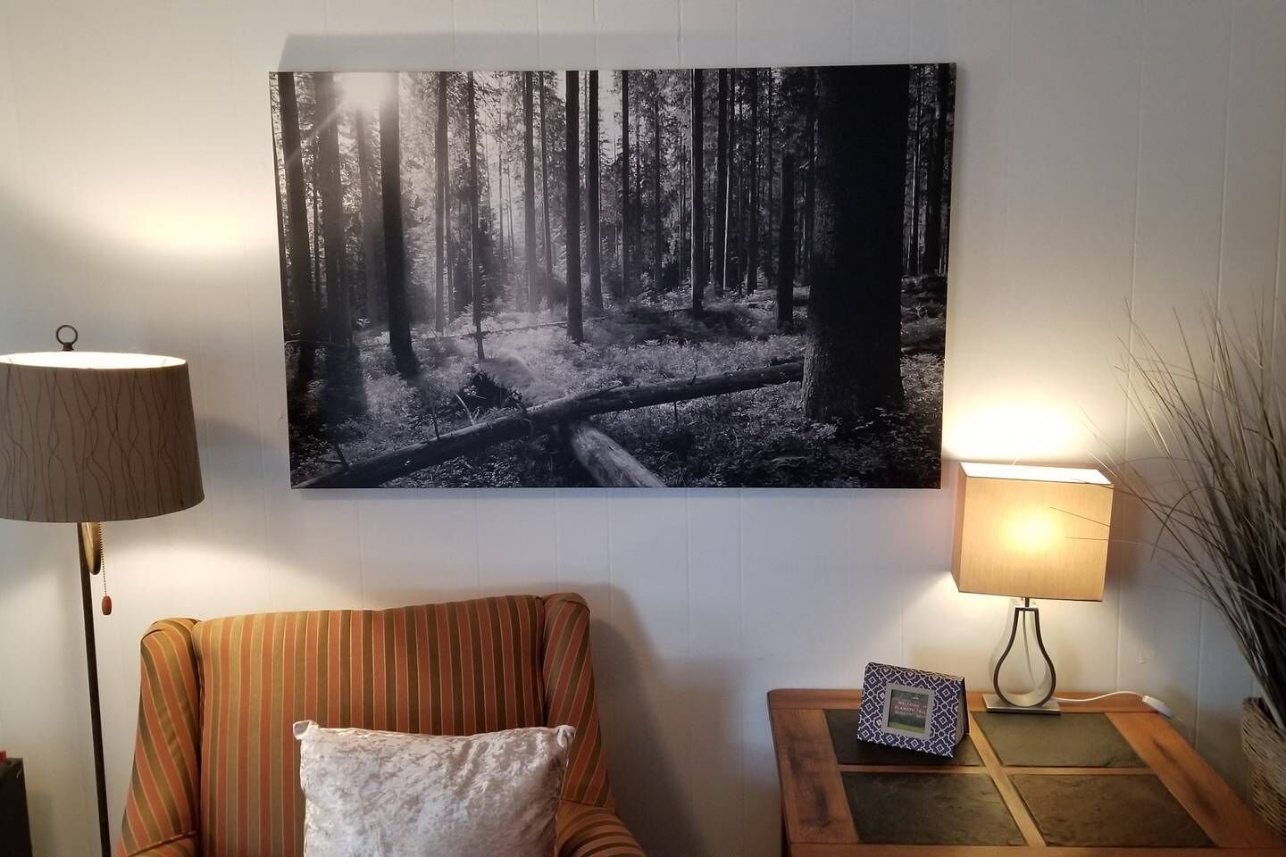 A room with a large forest-themed artwork on the wall, a striped sofa, a small side table with a lamp, and a decorative item on the table.