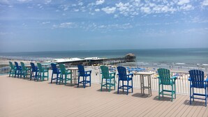 A deck with chairs and tables overlooking a beach with a pier and a thatched-roof structure.