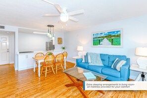 Un salon moderne avec un canapé bleu, une table à manger et des chaises en bois, un coin cuisine avec des meubles blancs et un ventilateur de plafond.