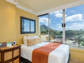 A hotel room with a large bed, a bedside table, a mirror, and a view of a mountainous landscape through large windows.