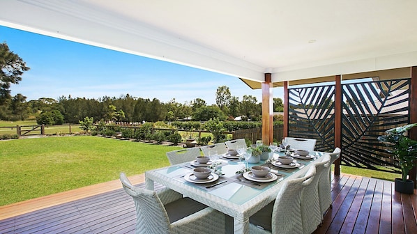 An outdoor dining area with a table set for four, surrounded by wicker chairs, on a wooden deck with a view of a landscaped garden and trees. - Best Cellar - The VIntage Holiday Houses (POKOLBIN)