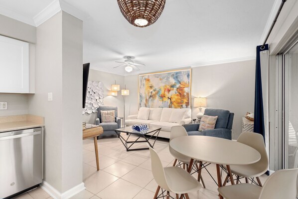 A modern living room with a white sofa, a blue armchair, and a dining table with chairs. There is a painting on the wall and a ceiling fan.