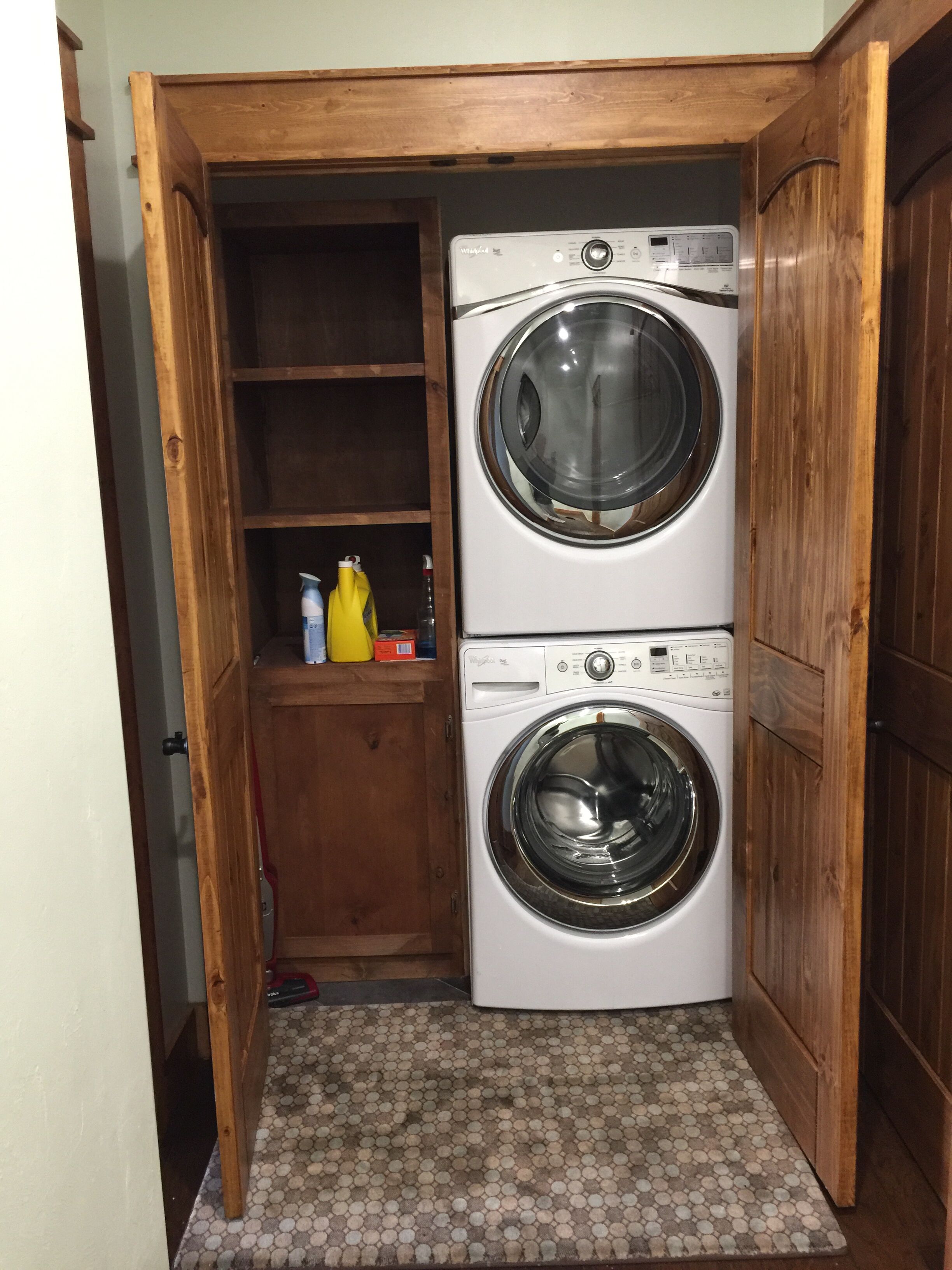 A laundry room with two stacked front-loading washing machines, wooden shelving, and a tiled floor.