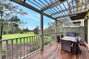 A covered outdoor area with a wooden deck, a grill, and a view of a green landscape.