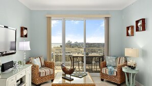 A spacious living room with two wicker sofas, a glass coffee table, and a television on a white cabinet. Large windows offer a view of the cityscape.