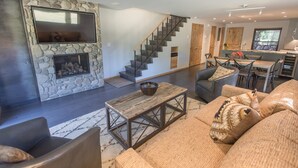 A modern living room with a stone fireplace, a large flat-screen TV, a staircase, and a dining area with wooden furniture. - Great 2 bed townhouse. This is the best location in Ketchum! (Ketchum)