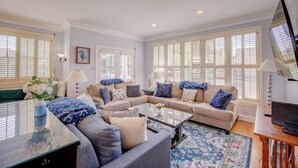 A spacious living room with a sectional sofa, a glass coffee table, and a large window with shutters. - Nantucket Charm | 280 ft from the Beach | Private Pool (Virginia Beach)