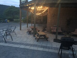 Un espace extérieur aménagé en patio, avec des tables et des chaises pour pique-nique, un barbecue et des guirlandes lumineuses.
