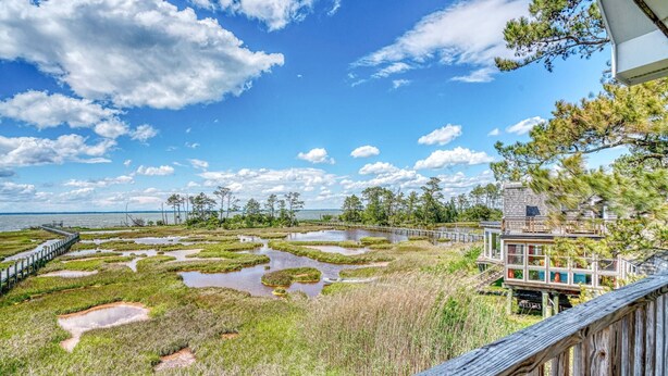 A coastal view with a wooden deck overlooking a marshy area with water bodies and a distant shoreline. - Castlebar | Luxury Salt Marsh Landing Waterfront w/ Private Pier, Bayfront Dock, Sunsets & Fireplace (Chincoteague Island)