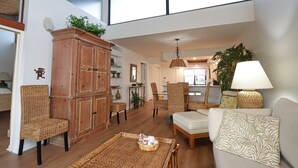 Un salon spacieux avec de hauts plafonds, une grande armoire en bois, des meubles en osier et une vue sur la cuisine.