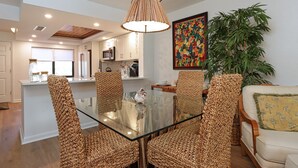 Un coin repas avec une table en verre et des chaises en osier, une cuisine en arrière-plan et une grande plante.