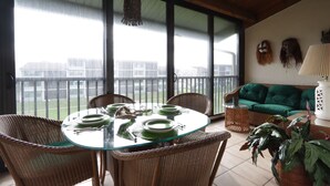 Un coin repas avec une table en verre dressée pour quatre personnes, entourée de chaises en osier. Un canapé vert et une lampe blanche sont visibles à l’arrière-plan.