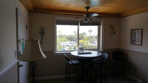 Un comedor con una mesa redonda, dos sillas, un ventilador de techo y una ventana que ofrece vistas a coches aparcados y árboles.