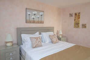 A neatly made bed with white and patterned pillows, a glass side table with a lamp, and a wall with a floral painting and two smaller artworks.