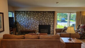 A living room with a stone fireplace, a sofa, a television, and a view of the outdoors. - Snowcreek Resort Vacation Rental 617 (Mammoth Lakes)