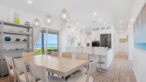 A modern kitchen with white cabinets, a stainless steel refrigerator, and a dining area with a wooden table and chairs. - New Latitude Beachfront Captiva Luxury Estate (Captiva)