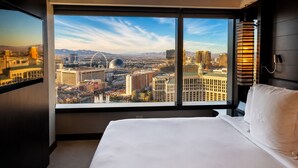 Habitación de hotel con una ventana grande que ofrece vistas a la ciudad, una cama y una mesita de noche con lámpara.