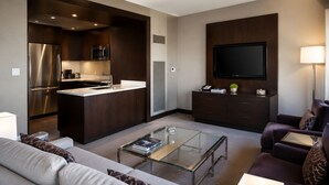 Una sala de estar moderna con una mesa de centro de vidrio, un sofá, un televisor, una zona de cocina con refrigerador y microondas, y un mueble con televisor de pantalla plana.