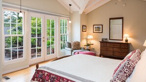 Une chambre avec un grand lit, une commode, une chaise et un bureau. Il y a deux fenêtres et une porte qui donne sur un espace extérieur.