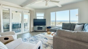 A spacious living room with a large flat-screen TV, a ceiling fan, and a balcony with a view of the ocean. - Completely Remodeled Direct Ocean View Colony Reef Condo 3401 (St Augustine)