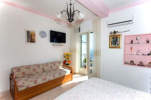 A bedroom with a wooden sofa, a bed, a TV, and a pink shelf with decorative items.