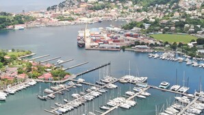 Ein Yachthafen mit zahlreichen Booten, ein Frachtschiff sowie Wohnhäuser und Hügel im Hintergrund.
