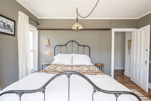 Une chambre à coucher avec un lit, des tables de chevet, un luminaire suspendu, une porte et une fenêtre avec des rideaux.