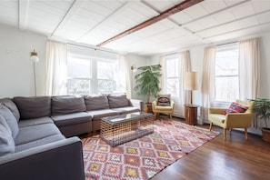 Un salon avec un canapé gris, un tapis à motifs, une petite table et une chaise avec un coussin.